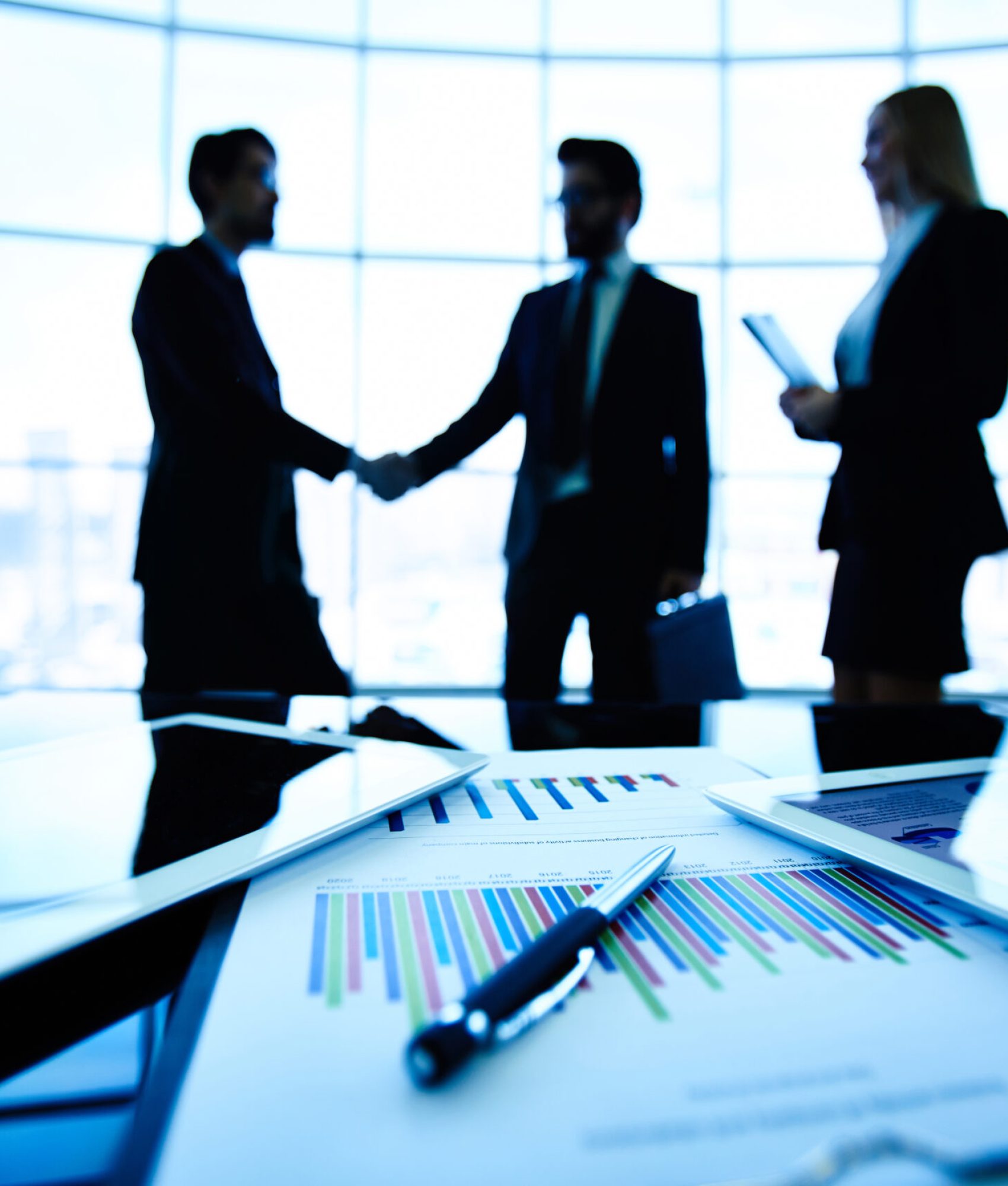 Technological devices, financial document with pen, glass of water at workplace on background of three business partners striking deal